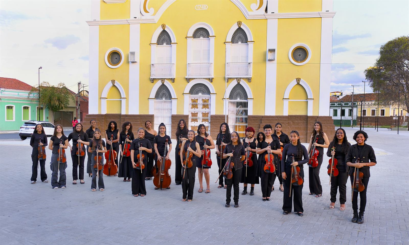 Alunas da Orquestra Aldo Parisot com seus instrumentos em frente a uma igreja amarela